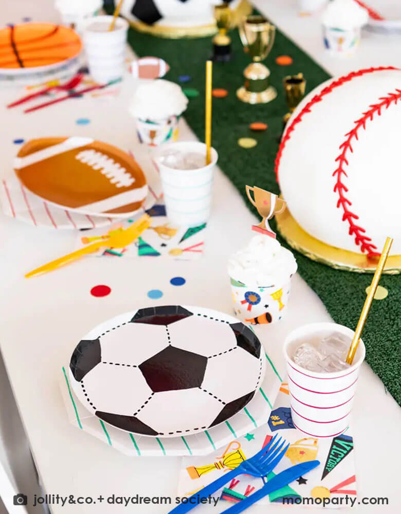 A fun party table filled with Momopartys's sports themed party tableware by Daydream Society including die-cut soccer ball shaped plates, football shaped plates, basketball shaped plates with mint and red striped plates and cups, plus some good sport large napkins featuring bright colors and fun illustrations of sports elements. In the center of the table there's a faux grass table runner and baseball shaped birthday cake, all makes great inspirations for kid's sport themed parties.