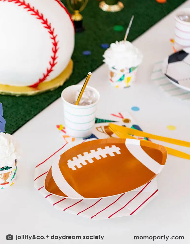 A fun party table filled with Momopartys's sports themed party tableware by Daydream Society including die-cut soccer ball shaped plates, football shaped plates, basketball shaped plates with mint and red striped plates and cups, plus some good sport large napkins featuring bright colors and fun illustrations of sports elements. In the center of the table there's a faux grass table runner and baseball shaped birthday cake, all makes great inspirations for kid's sport themed parties.