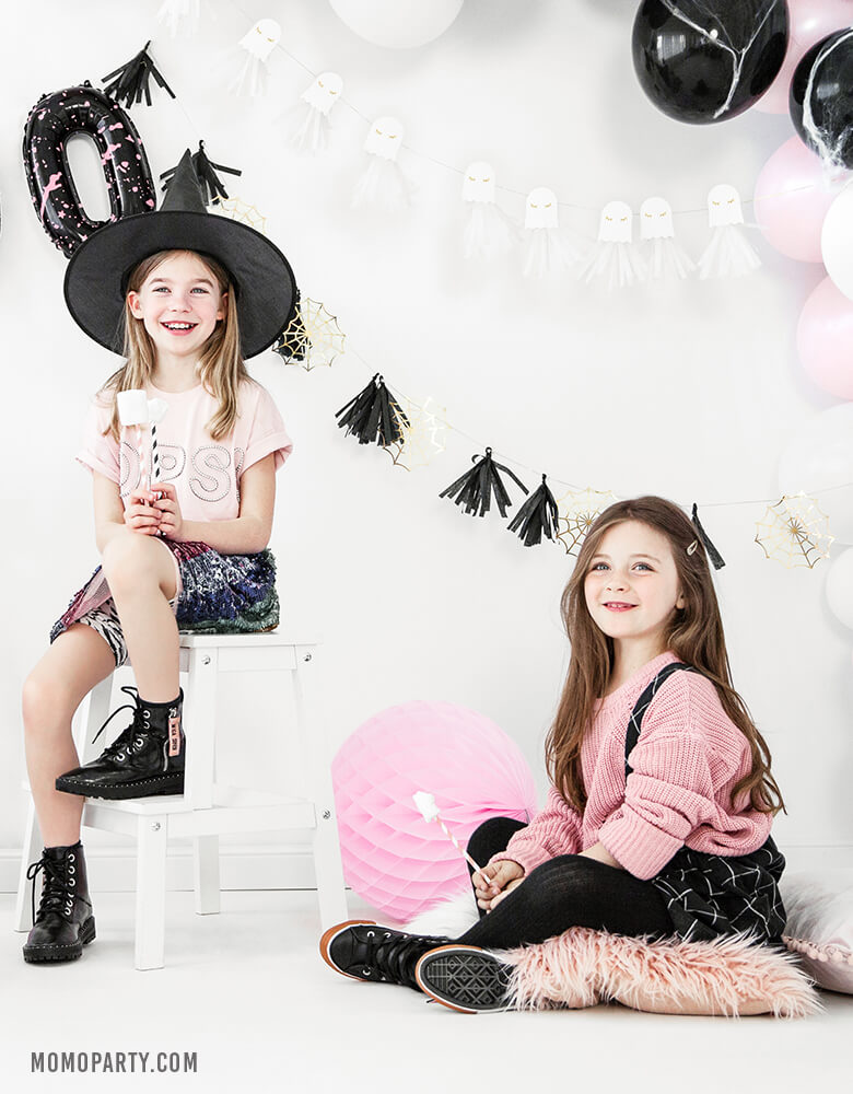 2 girls wearing witch hat, pink sweaters, seating in a room decorated with Party Deco golden spiderwebs and black tassels garland, ghost garland, pastel pink and black balloon garland, celebrating a Pink Halloween party. These cute halloween decorations also perfect for Kids modern spooky halloween party, trick-or-treating halloween party, nightmare before christmas party, witch themed party and all halloween related celebrations