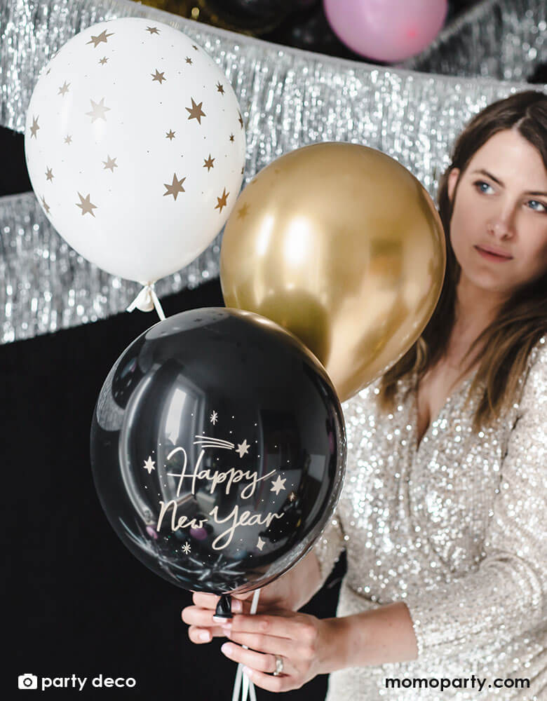 A lady in sequin party dress holding Momopartys's Happy New Year Latex Balloon Mix, set of 6 by Party Deco in the color of black, white and gold with Happy New Year message on it, a perfect decoration for a New Years Party celebration!