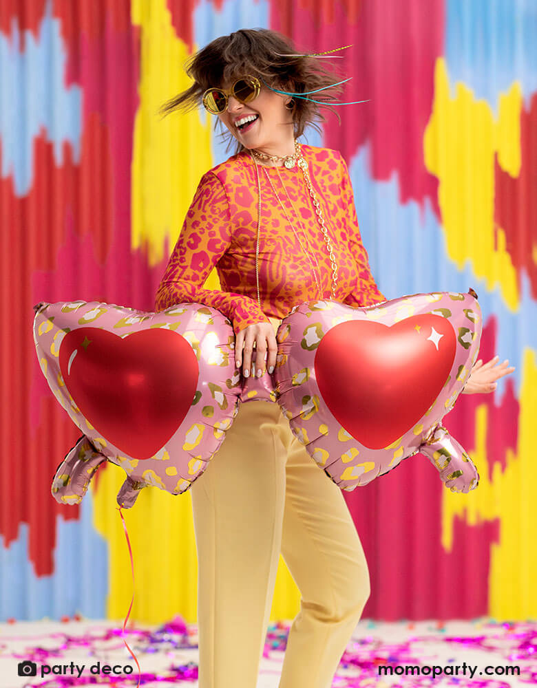 A young lady in her leopard shirt holding Momopartys's 30 inch heart shaped sunglasses foil balloon by Party Deco, With animal print design around the glasses in pink and red colors in her arms, standing in front of a colorful bright wall, celebrating love for Valentine's Day.