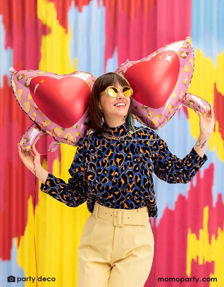 A young lady in her leopard shirt holding  Momopartys's 30 inch heart shaped sunglasses foil balloon by Party Deco, With animal print design around the glasses in pink and red colors on her shoulder, standing in front of a colorful bright wall, celebrating love  for Valentine's Day.