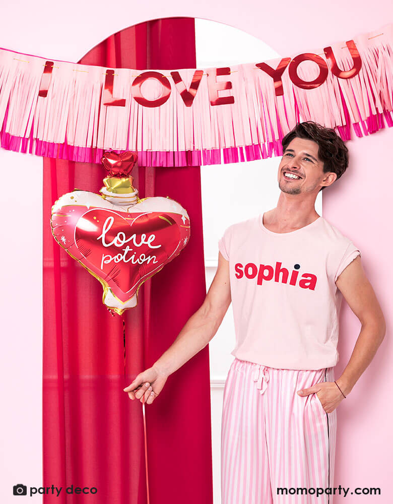 A man dressed in pink pajama holding Momopartys's love potion shaped foil balloon by Party Deco, standing in front of a door decorated with pink tissue-fringe banner with red type message of "I love you" - a perfect way to express your love on Valentine's Day.