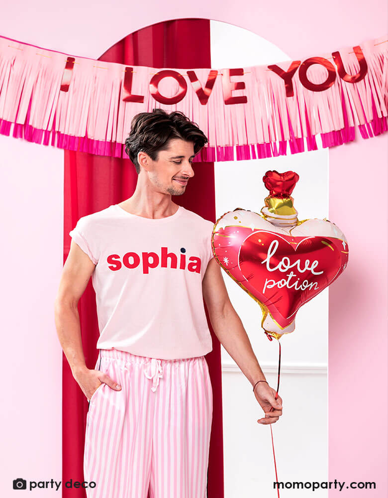 A man dressed in pink pajama holding Momopartys's love potion shaped foil balloon by Party Deco, standing in front of a door decorated with pink tissue-fringe banner with red type message of "I love you" - a perfect way to express your love on Valentine's Day.