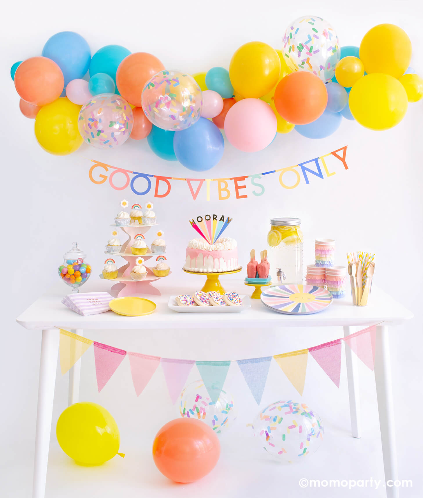 Momopartys Good Vibes Only Party Box Kit. Included Meri Meri Rainbow Sun Dinner Plates, Oh Happy Day Yellow Side Plates, Meri Meri Rainbow Sun Fringe Cups, Party Deco Good Vibes Napkins, Yellow Wooden Cutlery, Mixed Pastel Striped Straws, cupcake with rainbow and daisy candles. Meri Meri Hooray cake topper on a dripping cake, colorful gumballs, Lemonade on the table. Meri Meri Multicolor Bunting Banner spelled as "good vibes only" text , Good Vibes 6-foot- Balloon Garland Cloud as decoration.