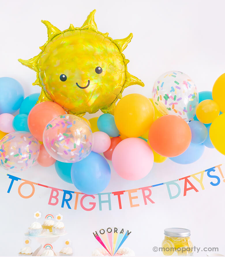 Momopartys Good Vibes Only Party Box Look, Backdrop details of Anagram Iridescent Sun Foil Mylar Balloon over a colorful blue, yellow, coral, goldenode mixed latex balloon garland with confetti latex balloons, and Meri Meri Multicolor Bunting Banner spelled as "To Brighter Days!" text