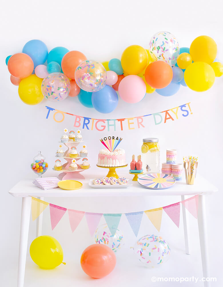 Momopartys Good Vibes Only Party Box Kit. Included Meri Meri Rainbow Sun Dinner Plates, Oh Happy Day Yellow Side Plates, Meri Meri Rainbow Sun Fringe Cups, Party Deco Good Vibes Napkins, Yellow Wooden Cutlery, Mixed Pastel Striped Straws, cupcake with rainbow and daisy candles. Meri Meri Hooray cake topper on a dripping cake, colorful gumballs, Lemonade on the table. Meri Meri Multicolor Bunting Banner spelled as "To brighter days" text , Good Vibes 6-foot- Balloon Garland Cloud as decoration.