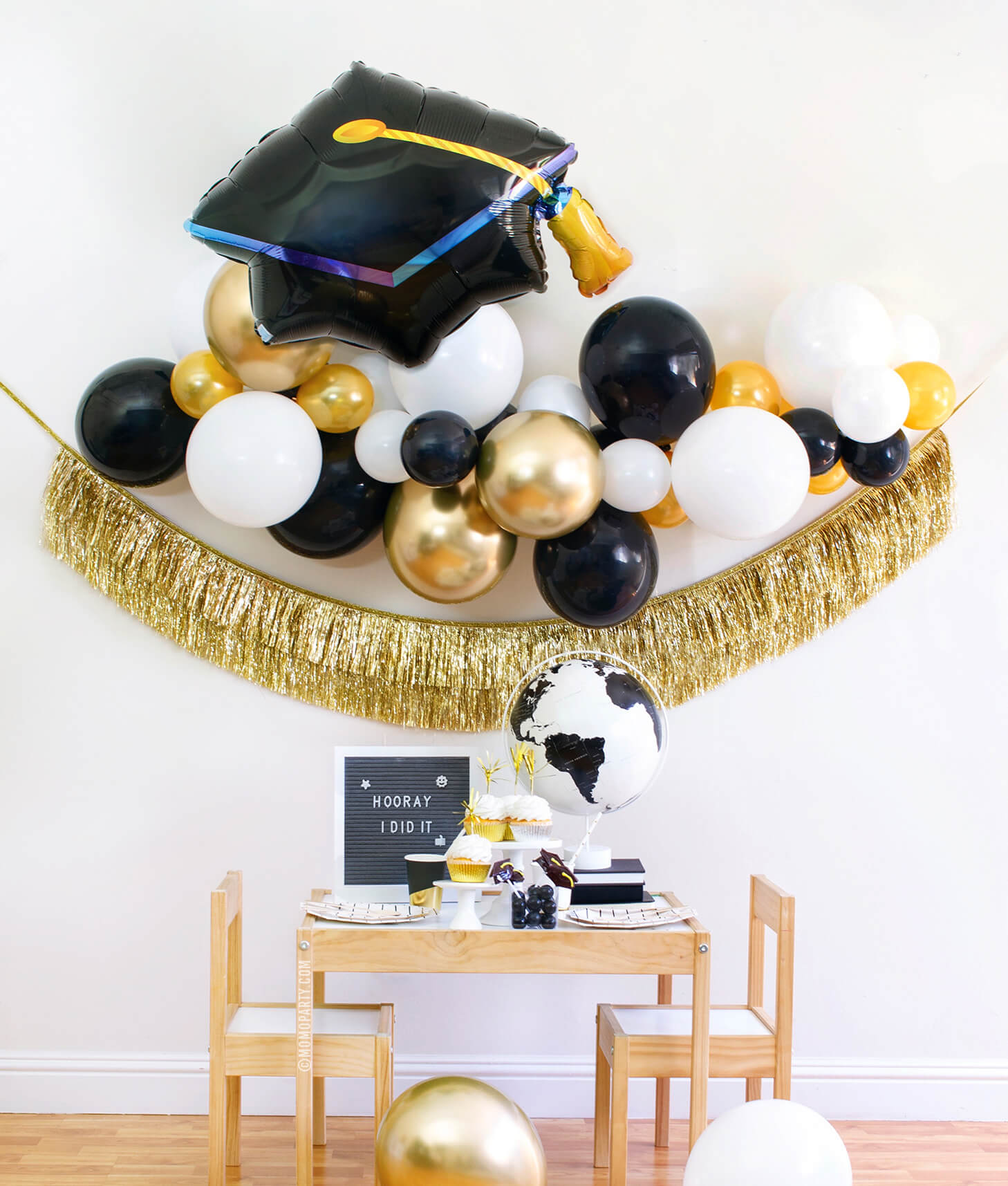 Momopartys modern graduation party at home box, come with black, gold and white Latex balloon garland, Black Graduation Cap Foil Balloon, Gold Tinsel Fringe Garland as backdrop decoration. Black and Gold stripe paper plates, stripe napkins, Assorted Gold Dipped Cups, b&w globe, letter board with "hooray i did it" sign, cupcakes, black gum balls on a kid table for a graduation celebration