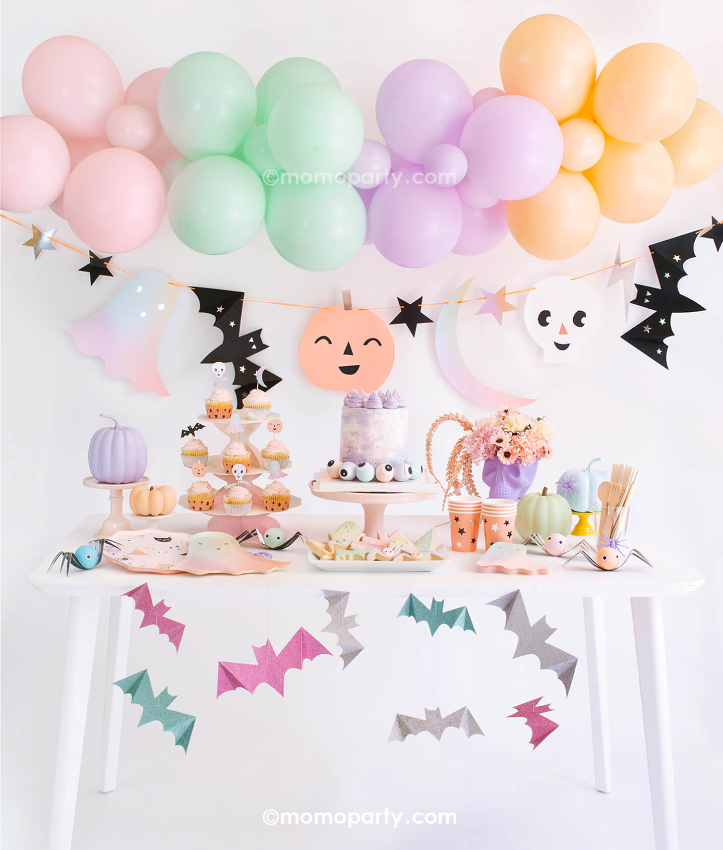 Momopartys Pastel Halloween Box Kit. Set up with Meri Meri Pastel Halloween Dinner Plates, Pastel Ombre Halloween Ghost Plates, Pastel Halloween Star Pattern Cups, Pastel Ombre Halloween Ghost Napkins, Wooden Cutlery, Iridescent Party Straws, Pastel Halloween Cupcake Kit  with 24 toppers, cake with eyeballs in the pink cake stand, skull head with flowers, spider surprise balls on the table. Pastel Halloween Glitter Hanging Bats, Pastel Balloon Cloud and Pastel Halloween Garland Set as decoration 