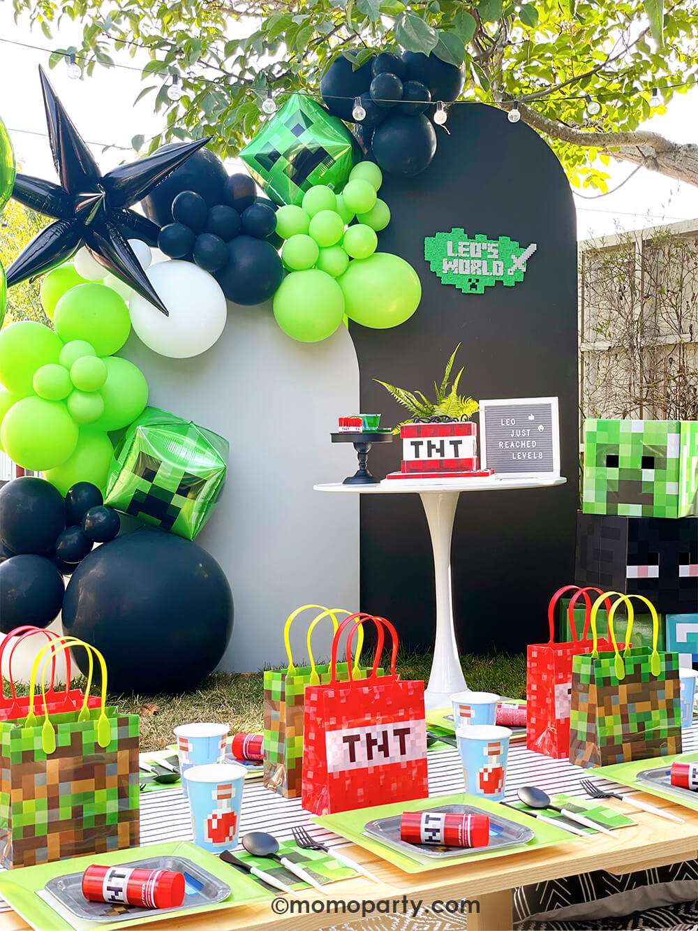 Kid's Minecraft themed Birthday Outdoor Party Ideas by Momopartys. Featuring Backdrop board in black and grey, decorated with minecraft themed color balloons in green, black, white, and creeper foil balloon, black starburst foil balloons. A TNT cake with letter board on the white round dessert table . In the front there is a kids picnic table filled with minecraft themed party plates with Minecraft Twist Poppers on top, cups, Creeper napkins and pixel TNT theme goodie bags on the black stripe table runner.