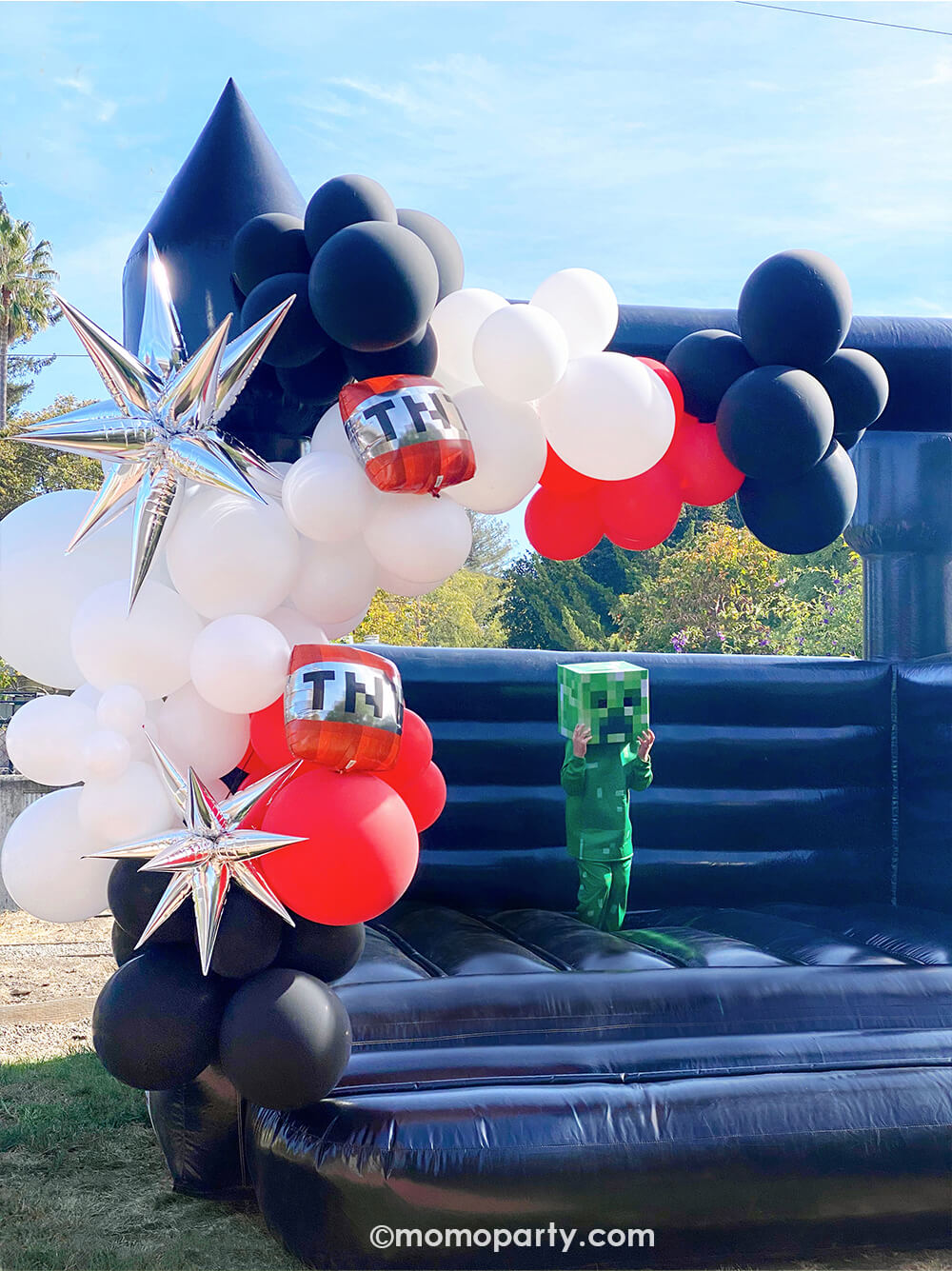 A modern Minecraft-themed birthday party by Momopartys. Featuring a giant black bouncing house adorned with black, white, and red latex balloons, 12 Point Silver Starburst Foil Balloons, and TNT foil balloons. A kid in a Creeper costume jumps with joy, making it a perfect and modern fun birthday party for boys.