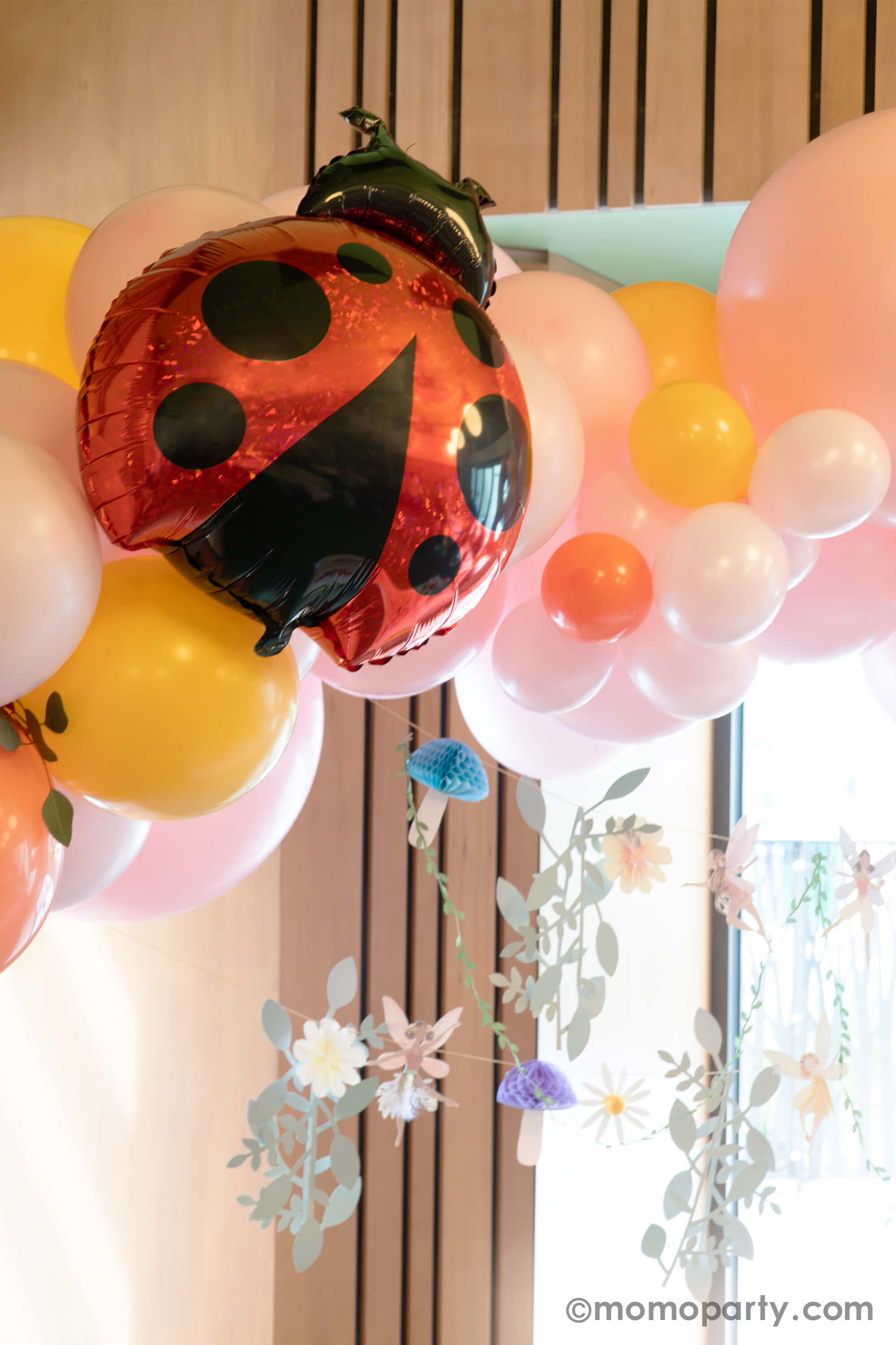 A spring fairy themed birthday party decoration featuring Momopartys's 27" ladybug shaped holographic foil balloon by Betallic Balloons on a spring inspired balloon garland in pastel pink, goldenrod, coral colors, with Momopartys's 6ft fairy garland by Meri Meri hung below it. All creates a beautiful decoration set up for a girl's birthday party in spring.
