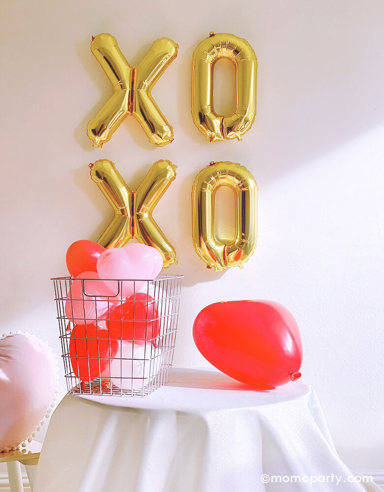 Momopartys Valentine's Day box, Party idea with Gold XOXO foil balloons as backdrop wall decoration, Heart Shaped Latex balloons in the silver basket, with a Red heart shaped latex balloon on the table, a pink heart shaped pillow on the side, a small home party set up for a Valentine's Day, Galentine's Day with friends, 