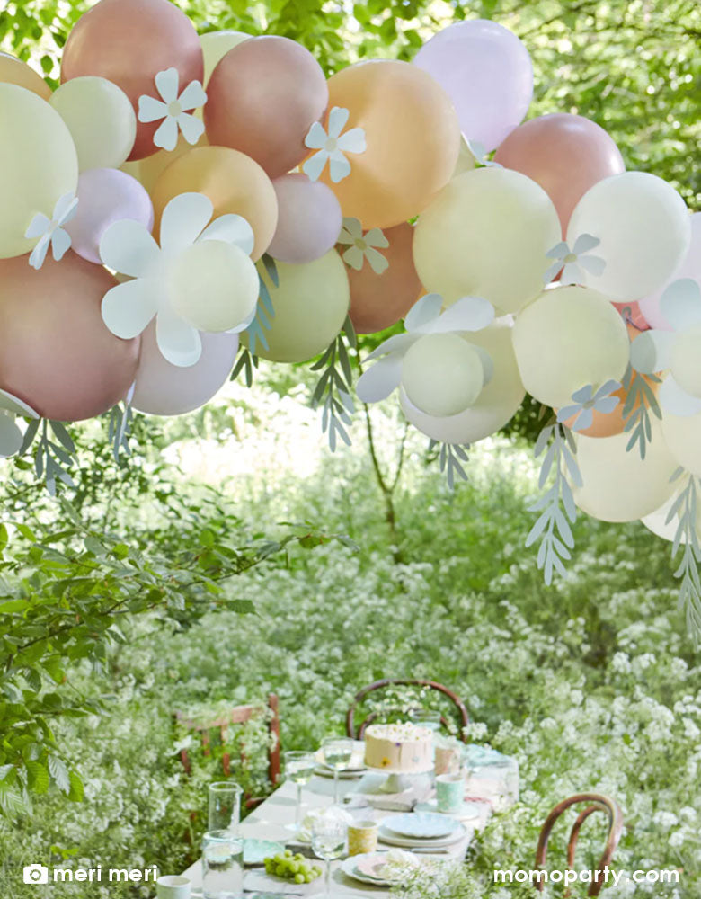 A beautiful party table set up in a field filled with wild daisy flowers. Above the party table it was Momopartys's Pastel Daisy Balloon garland by Meri Meri consisting 51 balloons in white, ivory, lilac, blush and rose gold with daisy shaped paper decorations and foliage shaped paper decorations on it. It makes a perfect decoration for a daisy themed birthday party or a spring inspired celebration.