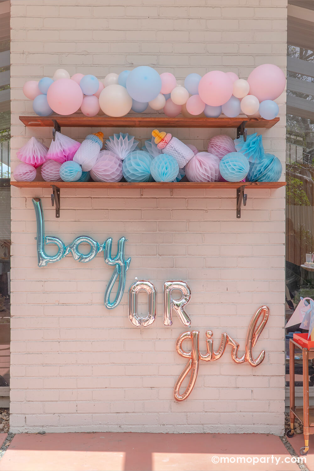 Boy or girl? A gender revel party by Momopartys, a brick wall decorated with boy or girl script foil letter balloons and a wall filled with honeycomb decorations in pink and blue and a boho pastel color balloon garland in baby pink and baby blue colors