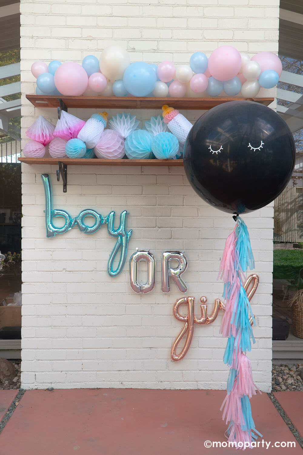Boy or girl? A gender revel party by Momopartys, a brick wall decorated with script foil letter balloons and a wall filled with honeycomb decorations in pink and blue and a boho pastel color balloon garland in baby pink and baby blue colors. In the front is the jumbo gender reveal confetti balloon decorated with pink and blue tassel, ready to be popped to reveal baby's gender!