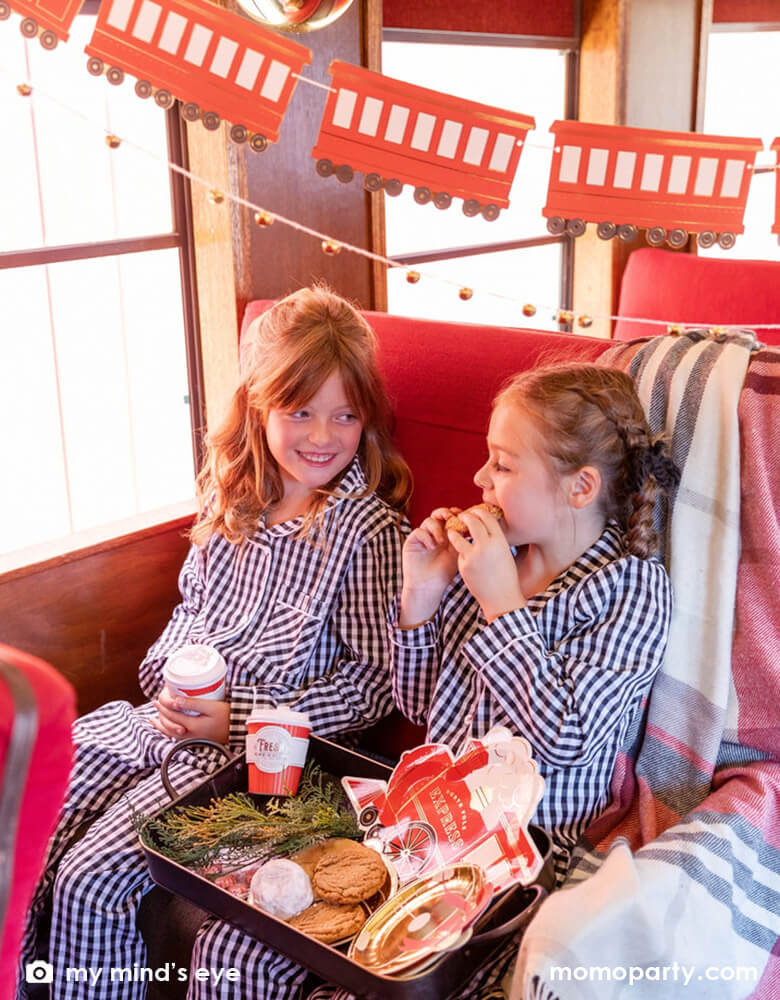 Two little girls in their Holiday pajamas enjoying a vintage tin tray filled with holly branches, holiday treats and cookies and Momopartys's North Pole Express train shaped plates, bell shaped side plates and polar express red 8 oz to-go cups by My Mind's Eye sitting on a vintage Polar Express train cabin decorated with Momopartys's North Pole Express train and bell garland set, a perfect idea for this Holiday season's celebration with family and kids.