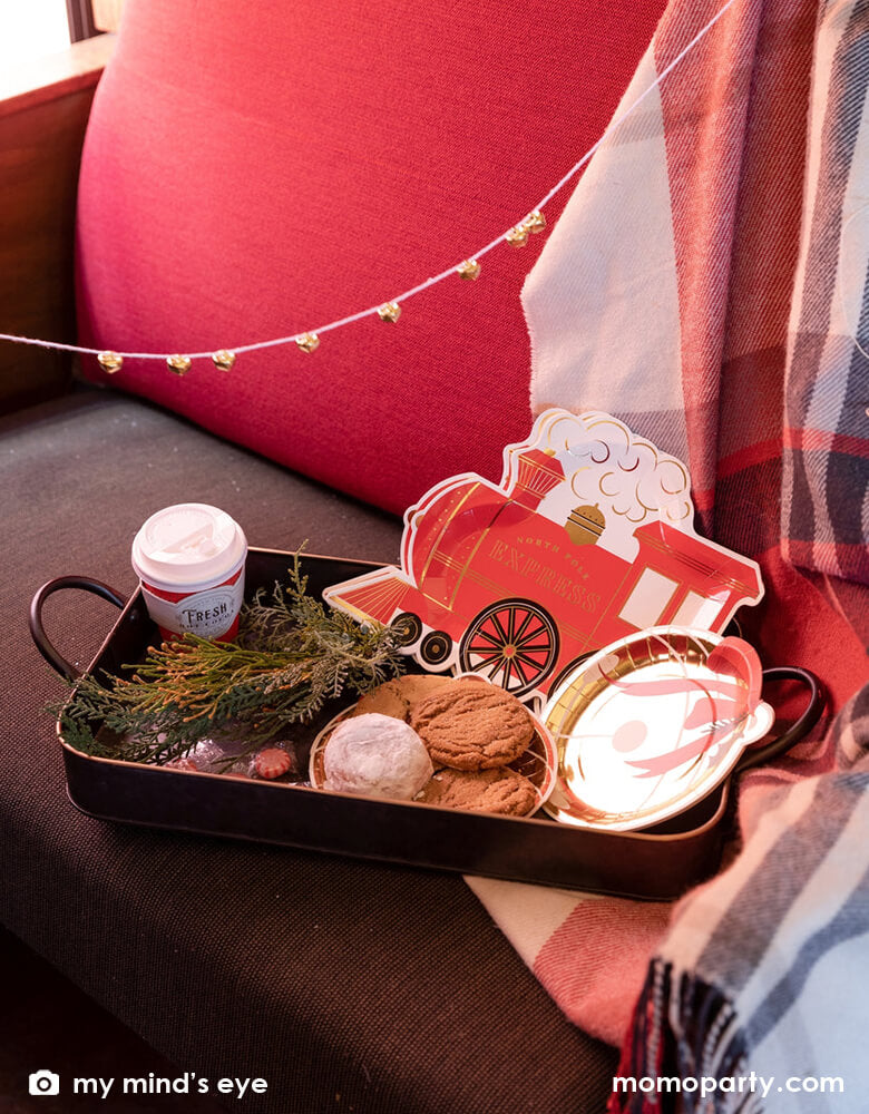 A vintage tin tray filled with holly branches,  holiday treats and cookies and Momopartys's North Pole Express train shaped plates, bell shaped side plates and polar express red 8 oz to-go cups by My Mind's Eye on a vintage Polar Express train cabin red leather seat, ready for a festive Holiday celebration for family and kids.
