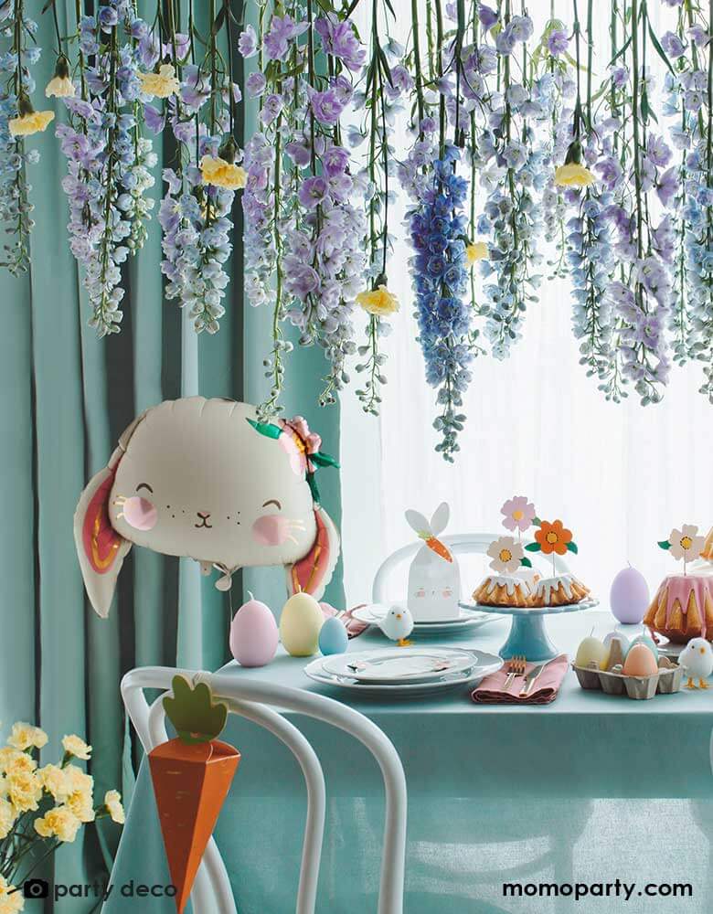A stunning table setup for an Easter celebration featuring Party Deco's 26" floral bunny foil balloon in front of beautiful floral arrangement hanging from above; on the table it's decorated with bunny themed napkins, carrot shaped goodie boxes and bunny shaped goodie bags and flower shaped cupcake toppers