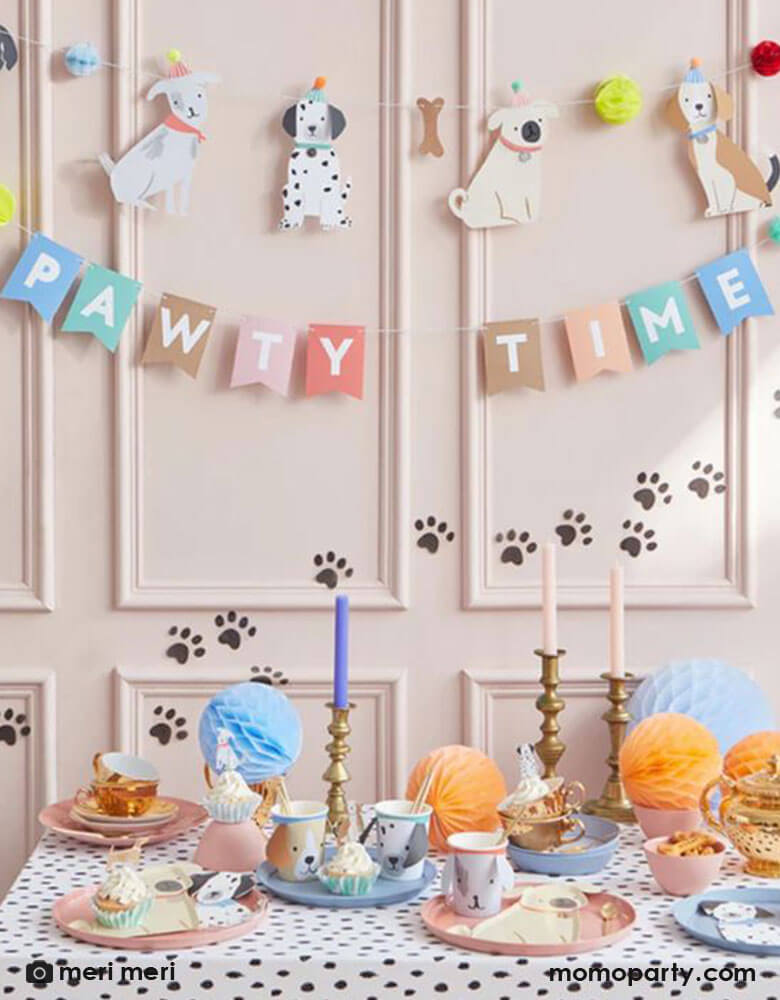 An adorable party set up for puppy dog themed birthday, featuring Momopartys's 6 ft Pawty Time party garland by Meri Meri, it features furry friends with embellished tails and pompom hats, bones and honeycomb balls. Drape it on the party table or on a wall for a delightful decoration. It's perfect for a dog lover's birthday party or a party for your family pet. In the front, a party table filled with puppy themed party supplies including dog party cups, pug shaped plates and Dalmatian shaped napkins.