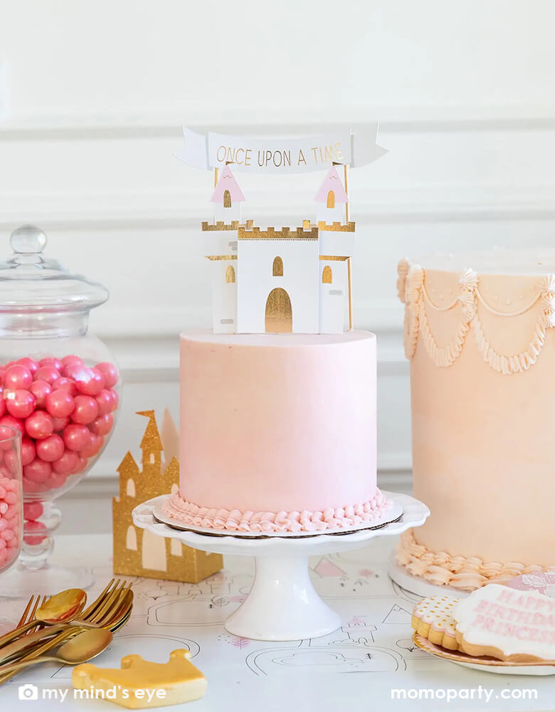 A gorgeous princess themed birthday cake decorated with My Mind's eye Cake topper set featuring castle pieces, mountains and a "Once Upon a Time" banner that will make any cake a the perfect addition to any princess party. Next to the cake, there are candy jars filled with pink gum balls, and some princess themed sugar cookies, a prefect dessert table inspo for girl's princess themed birthday party