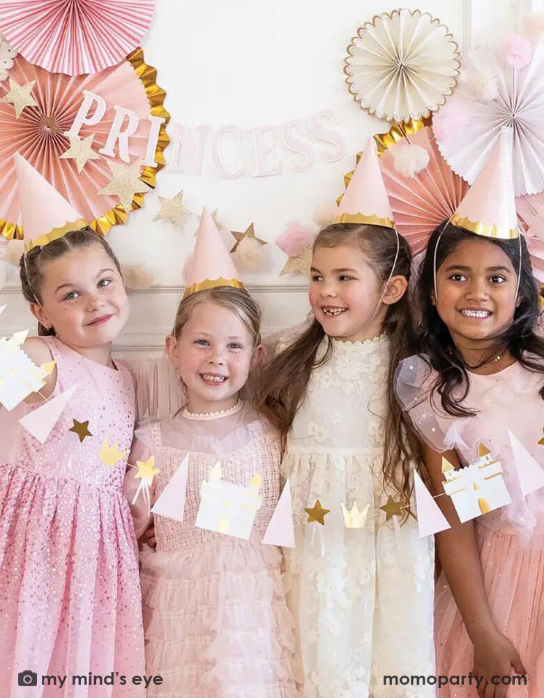 A group of 4 girls in their 6 or 7 years old dressed up as princesses with their tutu dresses and Momopartys's princess birthday hats, they're standing in front of a wall decorated with pink party fans with gold foil. Together they hold Momopartys's 5.5 ft princess icon banner featuring castles, crowns, and stars. A sweet look for girl's princess themed birthday party.