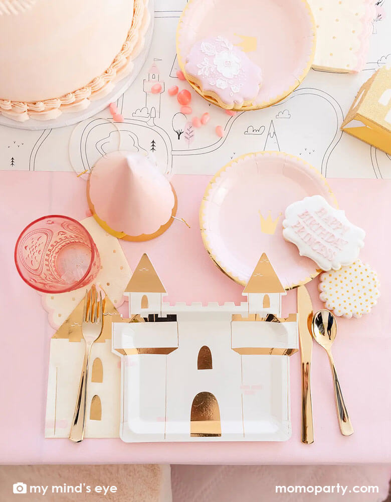 A beautiful tablescape from a girl's princess themed birthday party featuring Momopartys's princess castle shaped large plates, pink princess crown round side plate, turret shaped napkins, a pink princess birthday hat with tulle, in the middle it's a table runner featuring coloring princess fairytale illustrations, all in all makes a great inspiration for kid's princess themed birthday party.