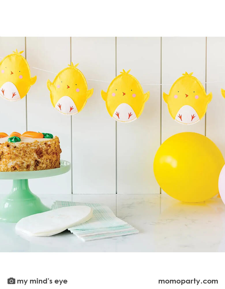 A kitchen counter decorated with Momopartys's 6 ft puffy felt chick banner by My Mind's Eye, with a carrot cake and easter egg shaped napkins and blue gingham guest towels, it creates a simply yet unique look for an Easter celebration.