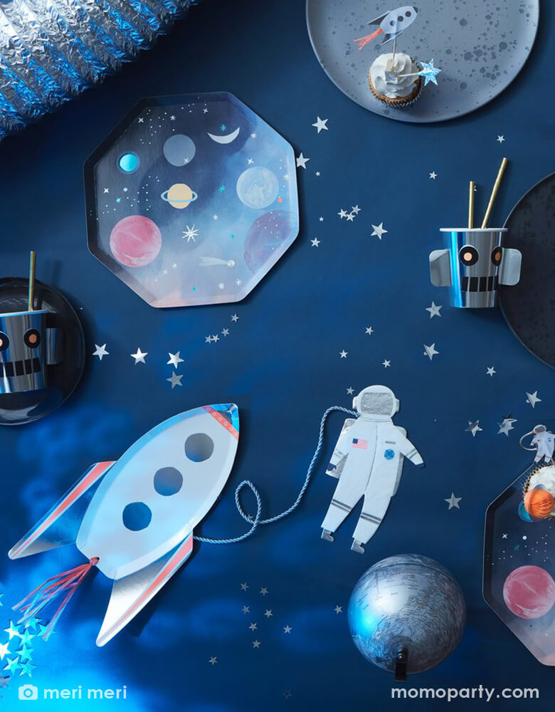 Top view of space themed party table, filled with Rocket Plates by Meri Meri, Space Dinner Plates, Robot Cup with gold straws, cupcakes decorated with space cupcake toppers on a moon plate, astronaut napkins and silver star confettis. These modern unique designed party supplies are perfect for a space themed birthday party, blast off birthday party, two the moon birthday party.