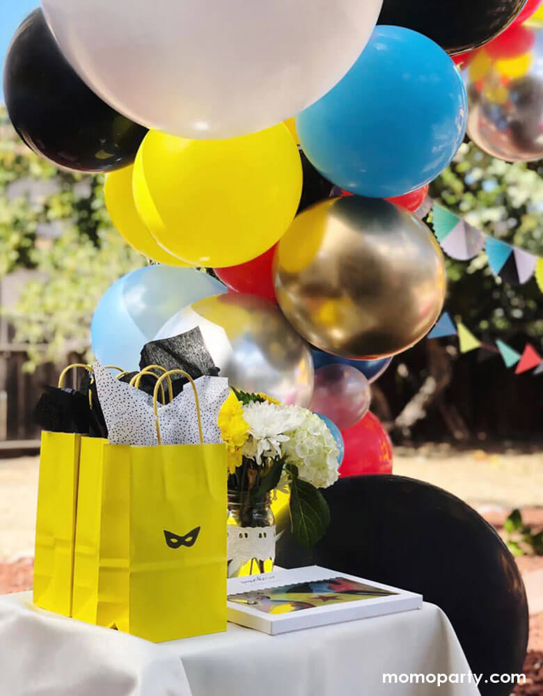 A kid's superhero party set up by Momopartys with a colorful balloon cloud in bright vibrant colors and superhero themed birthday banner, in the front of the table was a few goodie bags with a superhero mask stamp on it, a great way to great a superhero themed goodie bag in a modern way! 