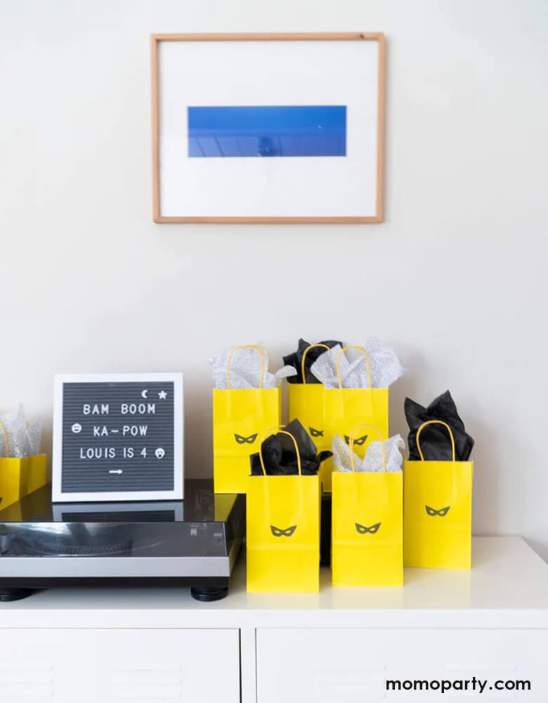 A table with yellow goodie bags decorated with Momopartys's superhero mask stamp, on the left is a modern letterboard spells out Bam Boom Ka-pow for a boy's birthday party celebration