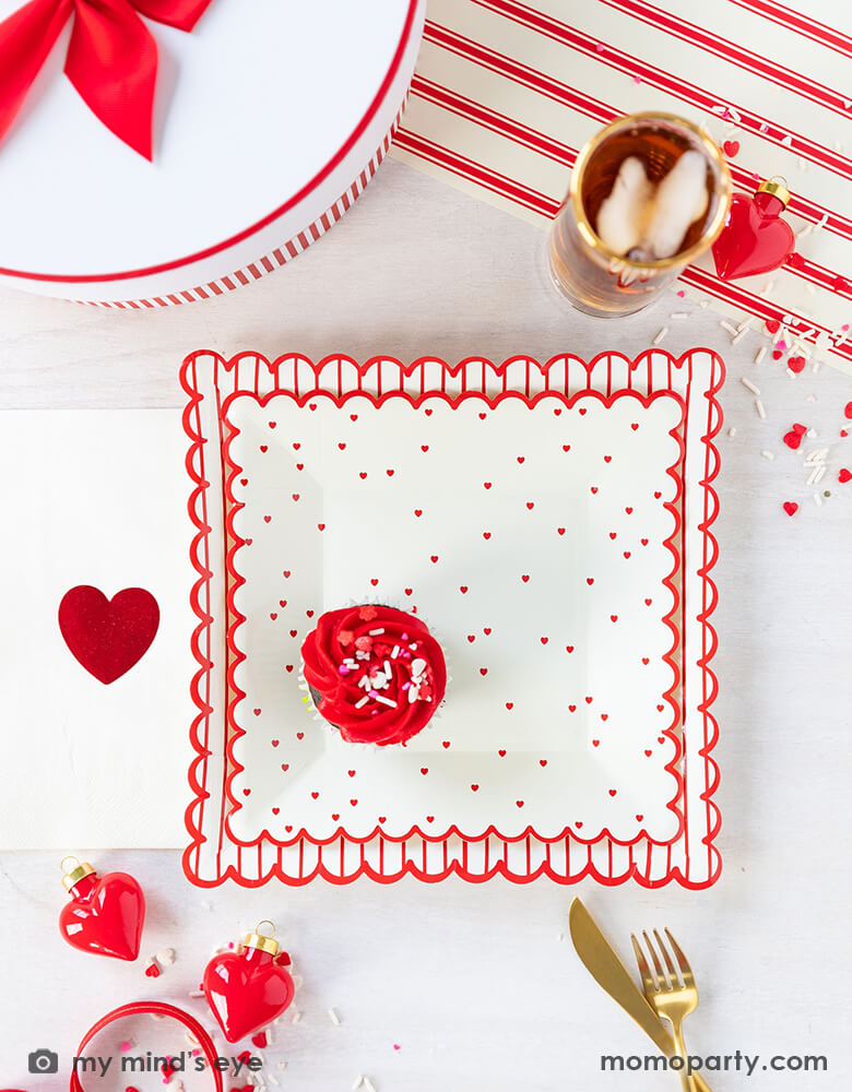 A sweet Valentine's Day celebration table filled with Momopartys's 8" square scattered red heart scallop plate by My Mind's Eye, Valentine Red Foil Heart Guest Napkins, heart shaped decoration, confetti, drinks and gold utensils over a Believe Christmas Red Striped Table Runner, for a sweet valentine's day party
