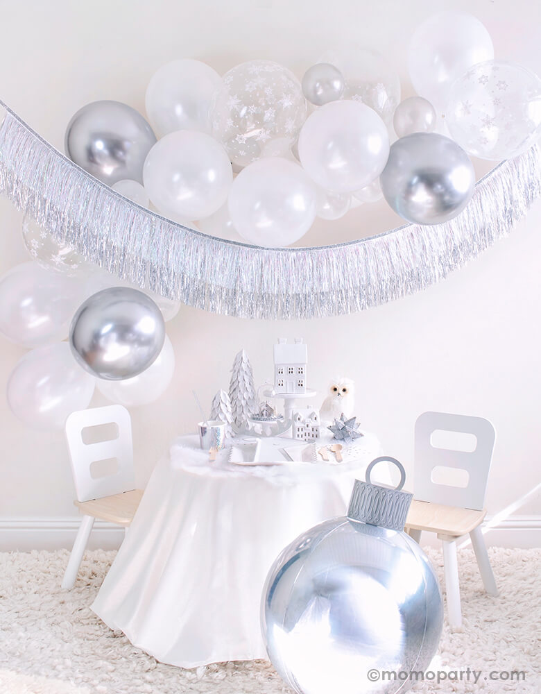 Momopartys 2020 Holiday White Christmas Holiday Box, Small party set up with White Scalloped Large Plates, Silver Sparkle Reindeer Plate, Silver Sparkle Tumbler Cup, Gleaming Large Napkin, Silver Wooden Cutlery and white trees, white house decor on the cake stand, Owl and silver star ornaments on the table, Silver Ornament Foil Balloon in the front. White, silver, snowflake mixed latex balloons cloud, and Silver Tinsel Fringe Garland as wall decoration for a simply modern gorgeous white Xmas party