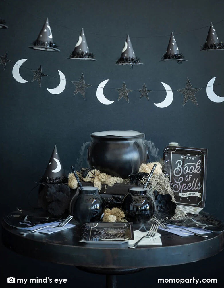 A Halloween witch themed party table set up with cauldron of witch brew and witch themed tableware including My Mind's eye book of spell shaped plates, witch hat shaped plates, witch candle napkins and witch hats, with moon and star  and witch hat shaped banner hung on above, all creates a spooky vibe for a frightening Halloween Eve